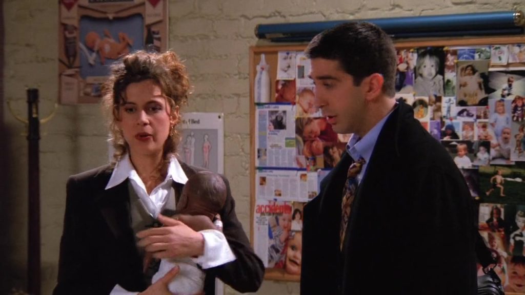 Ross and a woman in a parenting class holding a baby, discussing family dynamics in Friends S01E16.