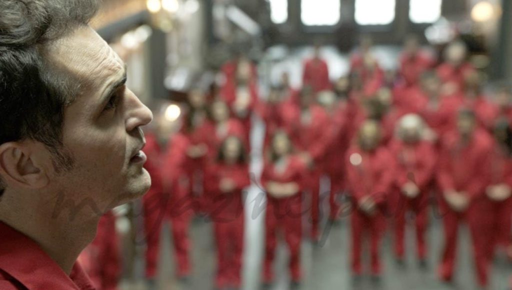  A tense crowd scene from Money Heist S01E06, showcasing hostages in red jumpsuits during the heist.