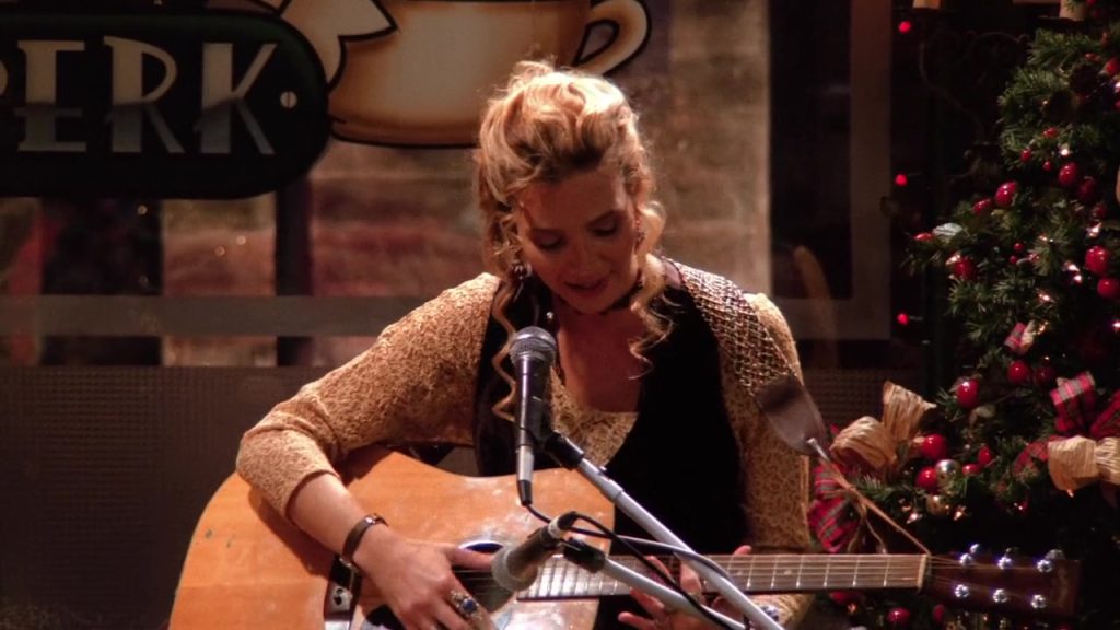 Phoebe Buffay playing guitar at Central Perk in Friends S01E10, showcasing her unique humor and style.