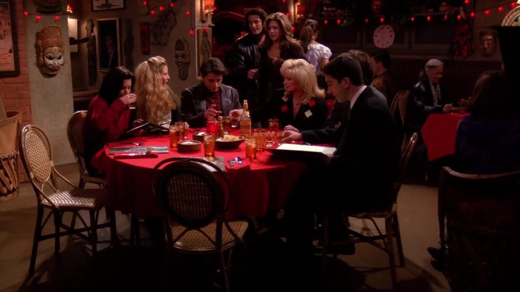 Friends characters sitting around a table in a restaurant, engaged in lively conversation.