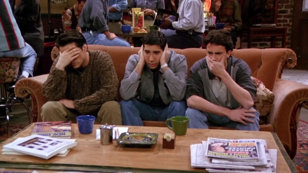 Ross, Chandler, and Joey sitting on the Central Perk couch covering their faces