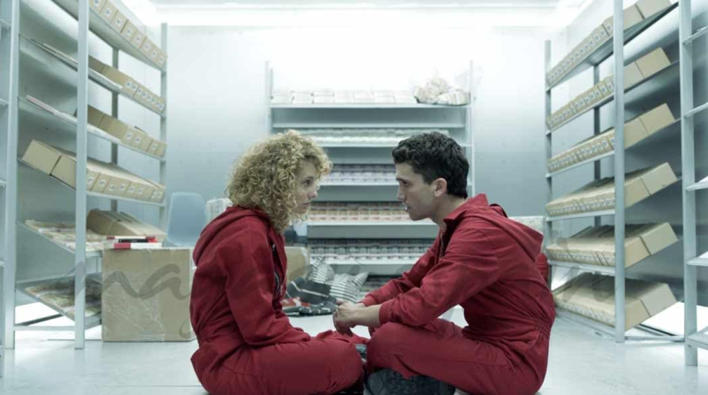 Two Money Heist characters sitting in red jumpsuits, engaging in an intense conversation.