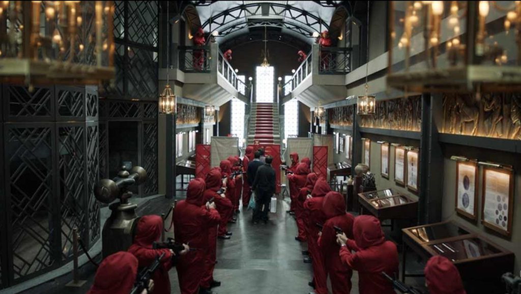 A grand hall with red-suited heist members aiming weapons at central figures.