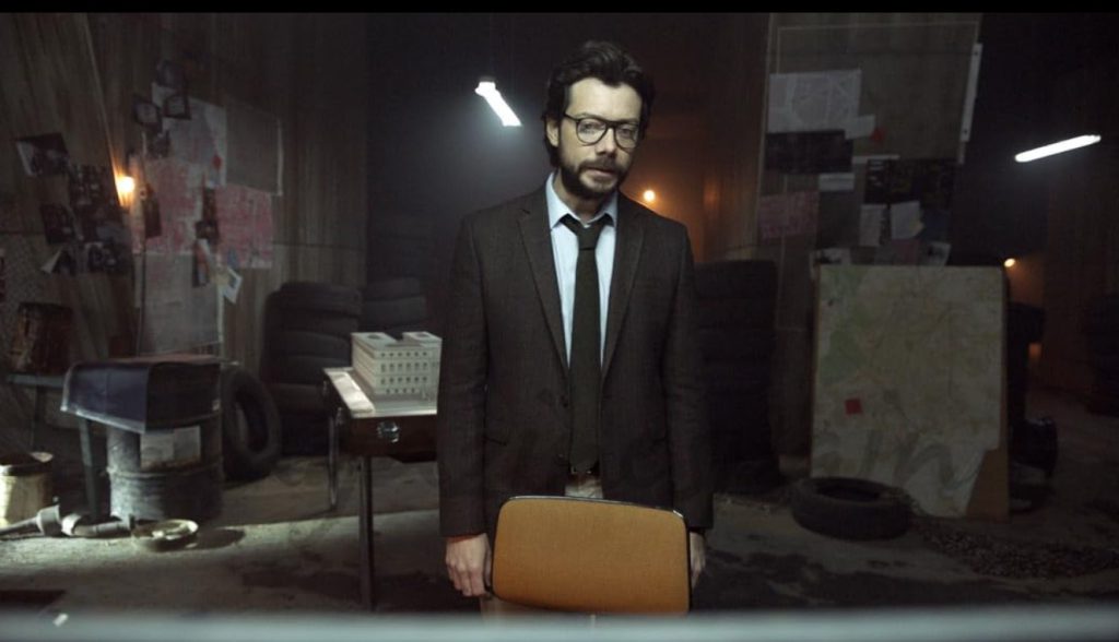 The Professor standing in a dimly lit planning room, surrounded by notes and heist layouts.