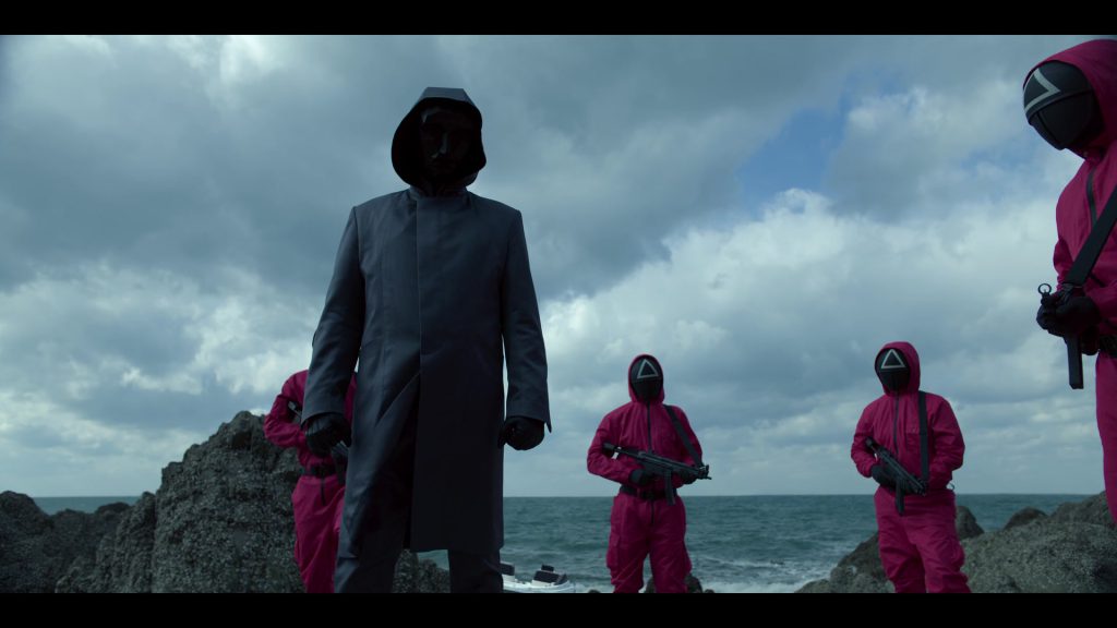 Squid Game guards in pink uniforms with weapons under a cloudy sky.