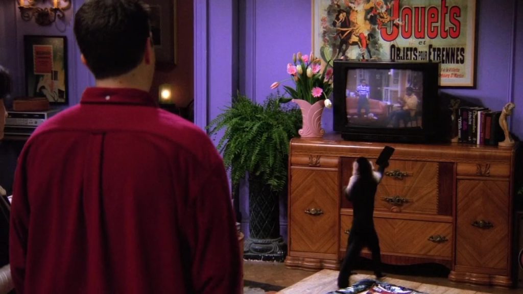 A black monkey holding a remote while facing a TV screen in the Friends S01E16 episode.