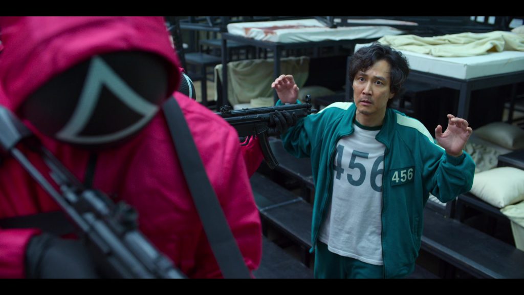 Squid Game character Gi-hun facing a guard pointing a gun, a powerful scene for movie vocabulary learning.