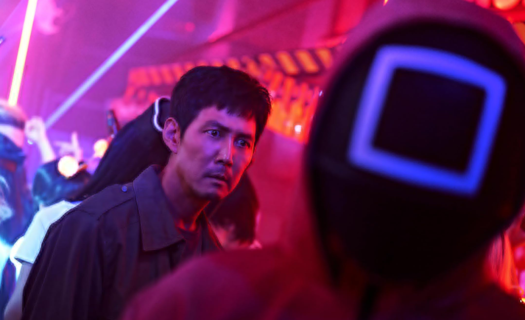 Film Vocabulary Builder with Squid Game – Language Learning with Netflix A tense scene from Squid Game S02E02 showing Gi-hun watching a masked guard during a Halloween party, perfect for language learners using a Film Vocabulary Builder.
