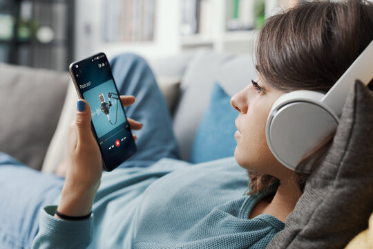 A young woman wearing white headphones, lying on a couch while listening to a language learning podcast on her smartphone.