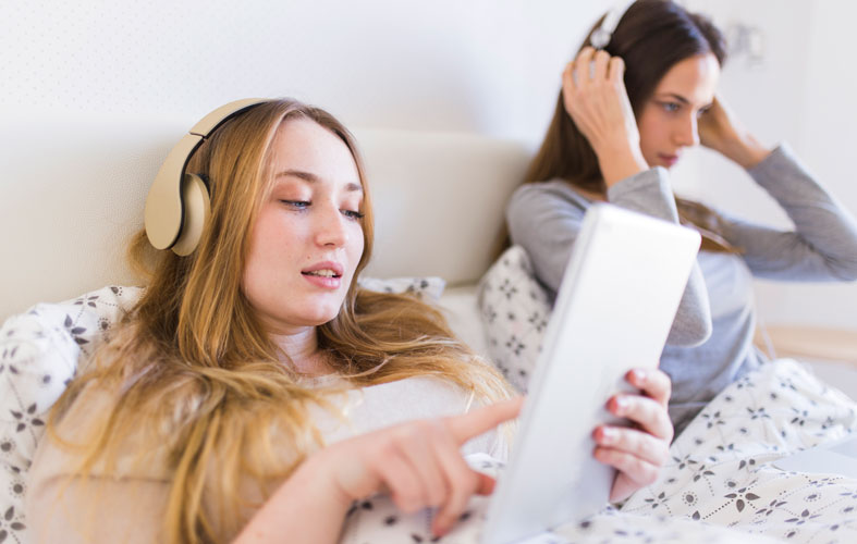 Two women lying in bed wearing headphones, listening to podcasts on Spotify