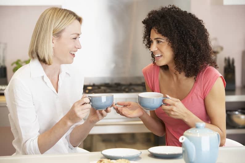 Two women smiling and enjoying tea in a cozy kitchen, symbolizing friendly conversations and language learning.